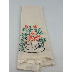 May include: Cream-colored kitchen towel with an embroidered design of a smiling teacup overflowing with orange flowers and green leaves. The teacup and saucer are outlined in black thread.