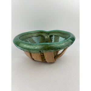 May include: A decorative bowl with a green glass rim and a woven, tan-colored base. The bowl's rim has a unique, heart-shaped design. The glass appears to be textured, and the bowl is set against a white background.