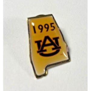 May include: A gold-colored pin in the shape of the state of Alabama. The pin features the number "1995" above the Auburn University logo in black. The background is a shade of orange.