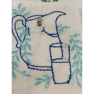 May include: White embroidered kitchen towel featuring a blue pitcher and glass design. The pitcher has a brown eye and is surrounded by blue and teal leaves. The glass has a white straw. The design is embroidered with blue thread.