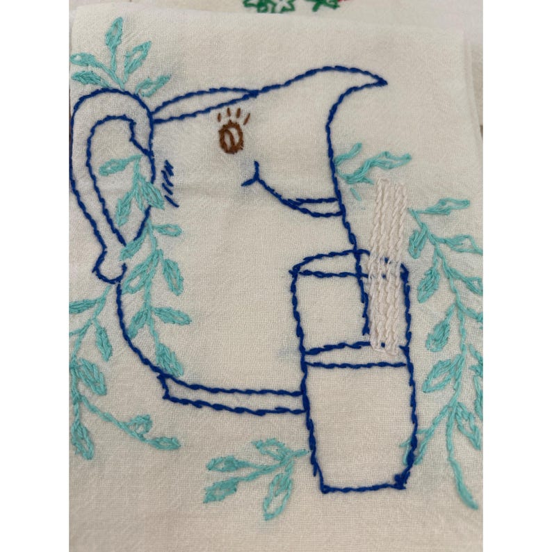 May include: White embroidered kitchen towel featuring a blue pitcher and glass design. The pitcher has a brown eye and is surrounded by blue and teal leaves. The glass has a white straw. The design is embroidered with blue thread.