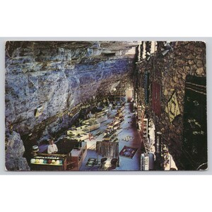 May include: Vintage postcard depicting a shop built into a cave. The shop features display cases with merchandise, stone walls, and a rock ceiling. Various rugs and decorative items hang on the walls. Several people are visible inside the shop.