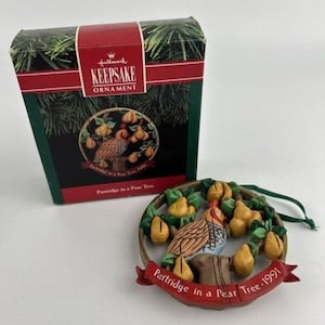 May include: A Hallmark Keepsake Christmas ornament from 1991, featuring a partridge in a pear tree design. The ornament is round with a brown, tan, and white bird surrounded by yellow pears and green leaves. The box is green and red.
