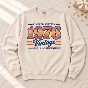 Könnte beinhalten: Ein cremefarbenes Sweatshirt mit Rundhalsausschnitt und einem Aufdruck mit dem Text LIMITED EDITION 1976 Vintage 50 JAHRE ALLE ORIGINALTEILE. Das Design enthält ein gestreiftes Muster im Used-Look in den Farben Rot, Orange und Gelb.