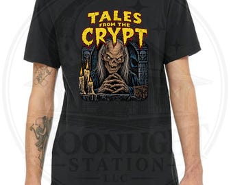 Può includere: Una t-shirt nera a maniche corte con una stampa grafica del logo Tales from the Crypt in testo giallo e rosso sopra un'illustrazione di una figura scheletrica con capelli lunghi, candele e archi in pietra in uno stile gotico scuro.