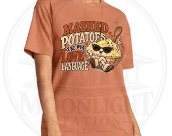 Può includere: Una t-shirt color arancio bruciato con la grafica di una ciotola di purè di patate che indossa occhiali da sole. Il testo recita MASHED POTATOES are my LOVE LANGUAGE. Il personaggio patata ha braccia, gambe e tiene un cucchiaio di legno.