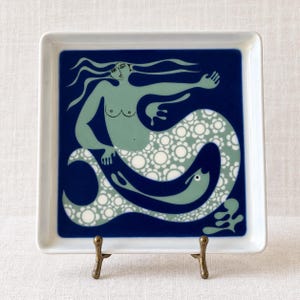 May include: A square white ceramic plate featuring a deep blue background with a central illustration of a light green mermaid. The mermaid has long flowing hair and a tail patterned with white floral circles, holding a small green fish.
