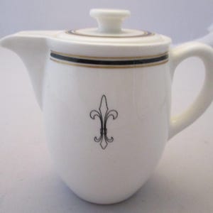 May include: White ceramic creamer with a lid, featuring a black and gold band around the top and a black fleur-de-lis design. The creamer has a handle and a spout for pouring. A classic piece for serving cream or milk.