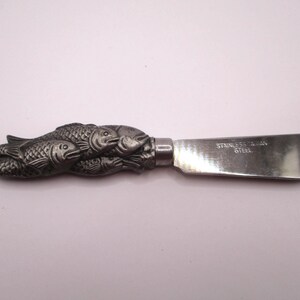May include: A stainless steel butter knife with a decorative handle featuring a cluster of fish. The blade is rectangular with rounded edges. The text "STAINLESS TAIWAN STEEL" is printed on the blade.