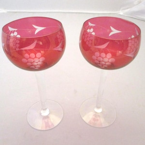 May include: Two red wine glasses with clear stems and bases. The glasses have a rounded bowl shape and are decorated with white etched grape and leaf designs. The glasses are on a white background.