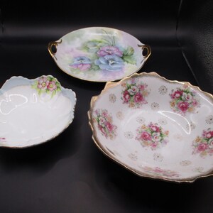 May include: Three porcelain dishes including a round plate with purple pansies and gold handles, a scalloped bowl with pink flowers and blue trim, and a larger oval bowl featuring pink rose clusters and gold gilded scalloped edges.