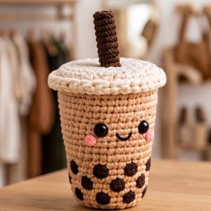 May include: A crocheted boba tea cup featuring a light tan body, a cream-colored lid, and a dark brown straw. The cup has a smiling face with black bead eyes, pink cheeks, and dark brown circular patches representing tapioca pearls.