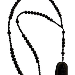 Puede incluir: Un collar largo compuesto por pequeñas cuentas redondas de color negro mate ensartadas, con un único colgante rectangular negro en la parte inferior. El colgante muestra un diseño en relieve texturizado en su superficie frontal.