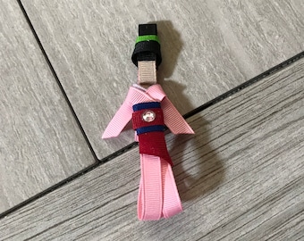 May include: A pink ribbon doll with a red and blue ribbon sash and a green and black hat. The doll is made of ribbon and has a rhinestone on the sash.