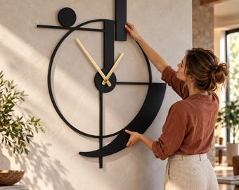 May include: A large wall clock featuring a black geometric frame with a circular outline, a vertical bar, and a small circle. The clock has two thin, gold-colored hands centered on the structure. The design is composed of matte black metal components arranged in an asymmetrical, abstract composition on a light-colored wall.