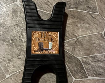 May include: A black plastic boot jack with a central rectangular label featuring the text JJ's BOOTJACK HIS N HERS and an illustration of two boots. The tool has a symmetrical shape with two U-shaped cutouts at the top and bottom ends.
