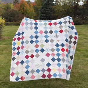 May include: A patchwork quilt featuring a grid of diamond-shaped fabric pieces in various colors and patterns, including red, blue, pink, yellow, and grey, separated by white fabric strips, creating a repeating geometric design throughout.
