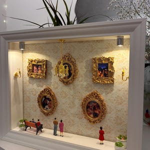 May include: A white shadow box frame containing a miniature room scene with beige patterned wallpaper, five ornate gold picture frames holding photos, a hanging gold chandelier, two wall sconces, a small bench, and four miniature figures.