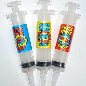 Puede incluir: Tres jeringas de plástico transparente con etiquetas que dicen "Super Power Shot" y tienen diferentes palabras de estilo cómic: "¡Zap!" "¡Pow!" y "¡Boom!"