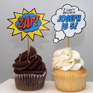 May include: Two cupcakes, one with chocolate frosting and the other with vanilla frosting. Each cupcake has a decorative topper. One topper says "ZAP!" in a comic book style. The other says "I CAN'T BELIEVE JOSEPH IS 5!"