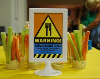May include: A yellow and black warning sign with a fork and knife graphic reads "WARNING! You are about to eat CICER ARIETINUM, DAUCUS CAROTA, APIUM GRAVEOLENS!"  The sign is on a white stand and there are four clear cups filled with celery, carrots, and hummus.