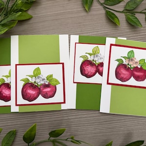 May include: Four rectangular greeting cards featuring a central illustration of red apples with green leaves and white blossoms. Each card has a light green vertical panel and a white border framed by a thin red line around the apple artwork.