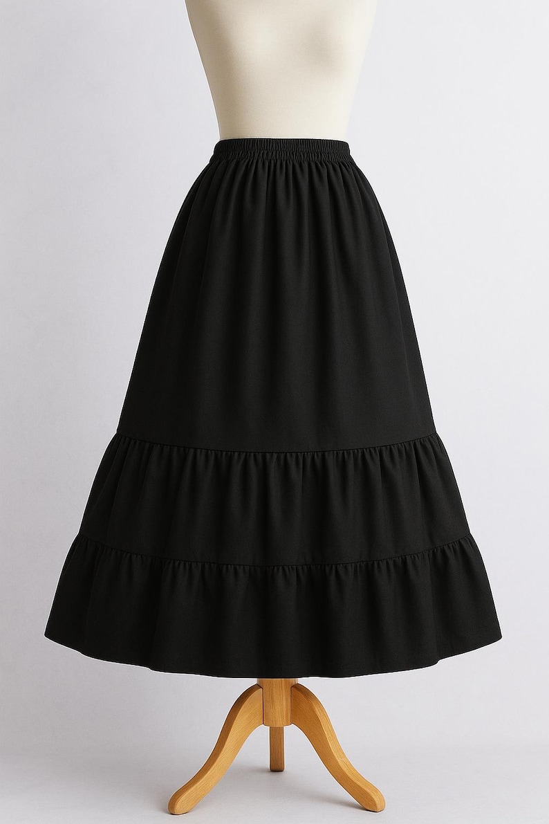 May include: A black, tiered skirt displayed on a mannequin. The skirt has an elastic waistband and a flowing, A-line silhouette. The skirt's hemline features two ruffled tiers, adding volume and a vintage aesthetic.