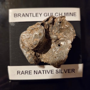 May include: A specimen of native silver from the Brantley Gulch Mine, featuring a rough, metallic gray, and textured surface. The mineral is displayed on a black box with white labels reading BRANTLEY GULCH MINE and RARE NATIVE SILVER.