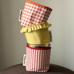 May include: Three stacked fabric jar covers with mango haus labels. The top cover is orange and white gingham, the middle is yellow plaid with a ruffled edge, and the bottom is red and white vertical stripes. Each features a white fabric tag.