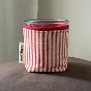 May include: A glass jar wrapped in a fabric sleeve featuring a red and white vertical striped pattern. The sleeve has a solid red top border and a small white fabric tag on the side with the black text mango haus printed vertically.