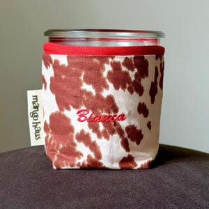 May include: A glass jar sleeve featuring a brown and white cow print pattern with a red fabric trim at the top. The name Bianca is embroidered in red script on the front. A white tag on the side displays the text mango haus in black.