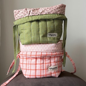 May include: Two stacked fabric storage baskets with tie closures. The top basket is green and white gingham with a pink floral lining. The bottom basket is pink with an orange grid pattern and pink polka dot lining. Both feature a mango haus label.