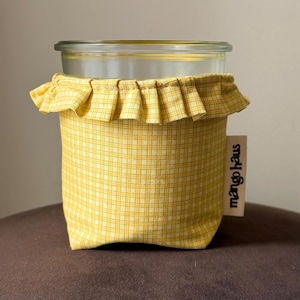 May include: A clear glass jar encased in a yellow and white checkered fabric sleeve with a ruffled top edge. A small beige fabric tag attached to the side features the black text mango haus printed vertically in a bold, sans-serif typeface.