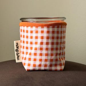 May include: A glass jar encased in a fabric sleeve featuring an orange and white gingham check pattern. The sleeve has a solid orange top hem and a cream-colored fabric tag on the side with the black text mango haus printed vertically.