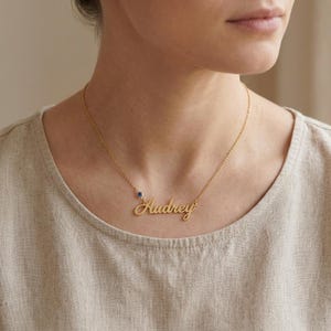May include: A gold-toned necklace featuring a delicate chain and a script-style nameplate pendant that reads Audrey. A small, round, dark blue gemstone is set into the chain just to the left of the nameplate, adding a subtle accent.