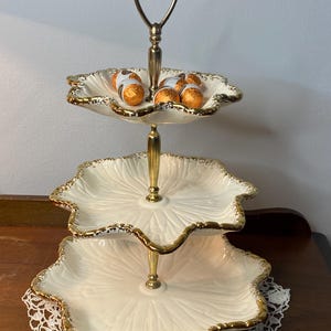 May include: A three-tiered serving stand featuring cream-colored ceramic plates with scalloped edges and gold-painted rims. The central support rod is made of gold-toned metal with a loop handle at the top. Several small, round, orange-wrapped chocolates sit on the top tier.