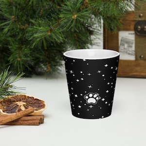 Puede incluir: Un vaso de chupito negro con un gráfico de huella blanca rodeado por un patrón de pequeñas estrellas y puntos blancos. El interior del vaso es blanco y la superficie exterior es lisa con una forma cilíndrica cónica.