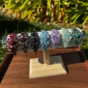 May include: A row of seven macrame bracelets displayed on a cylindrical stand. Each bracelet features intricate knotted cord patterns, small silver-toned beads, and a central gemstone in red, dark brown, black, light blue, lavender, mint, and green.