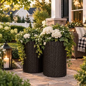 May include: Two tall, cylindrical dark brown woven planters filled with lush green ivy and clusters of white hydrangea flowers. The planters have a textured, basket-like appearance and are positioned on a stone patio surface.