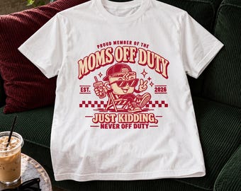 Può includere: Una t-shirt bianca con la grafica di una tazza di caffè con occhiali da sole e cappellino, seduta su una sedia a sdraio. Il testo recita: PROUD MEMBER OF THE MOMS OFF DUTY EST. 2026 JUST KIDDING, NEVER OFF DUTY in rosso e crema.