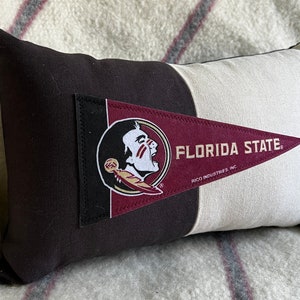 May include: A black and white pillow with a Florida State Seminoles pennant sewn on. The pennant is maroon and gold with the Seminole head logo and the words "Florida State" in gold lettering. The pennant also has the words "RICO INDUSTRIES, INC." in small white lettering.