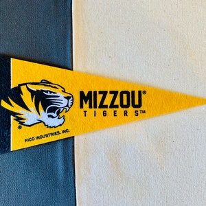 Pode incluir: Um estandarte amarelo com uma cabeça de tigre preta e o texto "MIZZOU TIGERS" em preto. O estandarte é feito de feltro e tem uma borda preta. O texto "RICO INDUSTRIES, INC." é impresso em preto na parte inferior do estandarte.