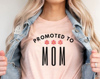 May include: A heather peach t-shirt with the text "Promoted to Mom" in black with three pink flowers above the text.