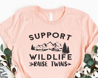 May include: A pink t-shirt with the text "Support Wildlife Raise Twins" in black. The text is surrounded by a graphic of mountains, trees, and a lake.