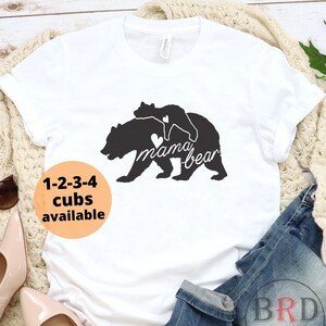 May include: White t-shirt with a black graphic of a mama bear and cub, with the text "mama bear" and a heart. The shirt also has the text "1-2-3-4 cubs available" in an orange circle. The word "WHITE" is at the bottom.