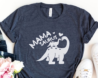 Peut inclure: Un t-shirt bleu marine chiné avec un motif blanc représentant une maman dinosaure et un bébé dinosaure. Le texte "Mama Saurus" est écrit au-dessus des dinosaures.