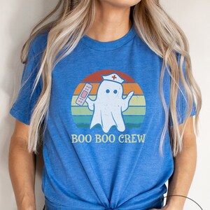 May include: A heather true royal blue t-shirt featuring a ghost wearing a nurse's hat and holding a bandage. The ghost is set against a retro sunset design with the words "BOO BOO CREW".