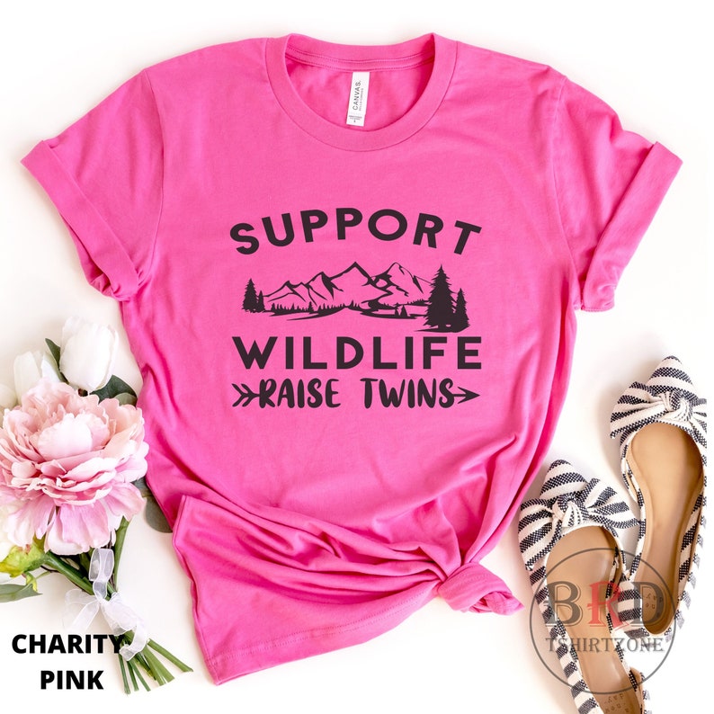 Puede incluir: Camiseta rosa con un gr&aacute;fico negro que dice "Apoyar la vida silvestre - Criar gemelos" con una cordillera y &aacute;rboles en el fondo.