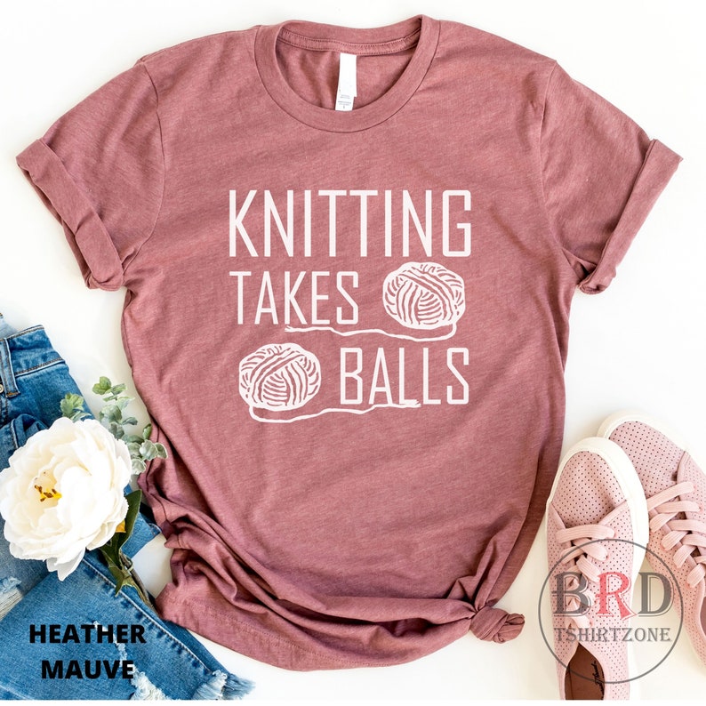 May include: A heather mauve t-shirt with the text "Knitting Takes Balls" and a graphic of two yarn balls.