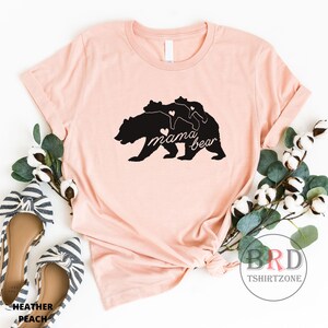 May include: Heather peach-colored t-shirt featuring a black graphic of a mama bear with two cubs and the words "mama bear". The design includes heart accents. The shirt is styled with flats and greenery.
